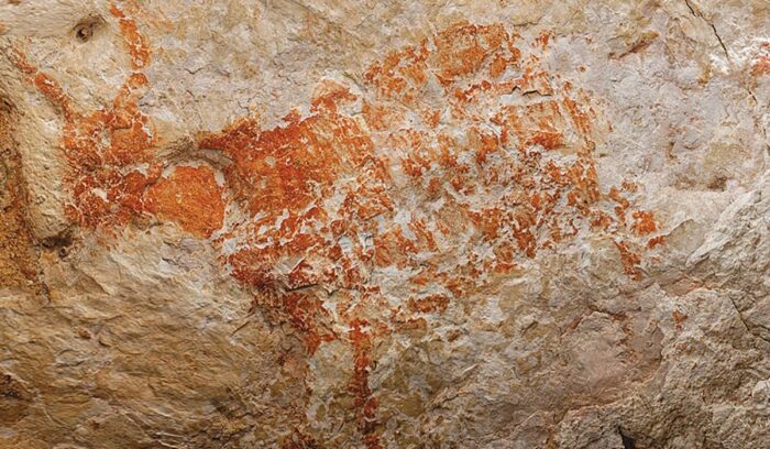 A faded orange hand stencil on a dark limestone cave wall for an article about Sulawesi cave art