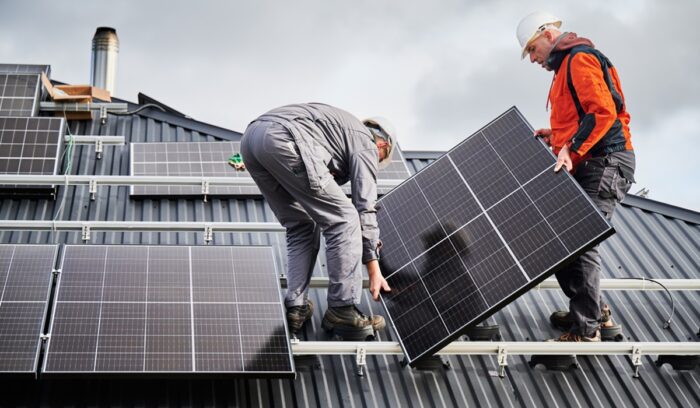 Technicians carrying photovoltaic solar module while installing solar panel system on roof of house, for article on Solar for All grants
